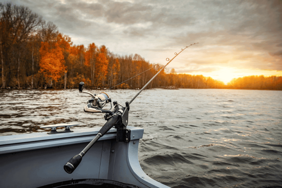 Image of a boat on a lake with a fishing pole cast off the stern. Leaves on the trees in the background indicate it is fall.