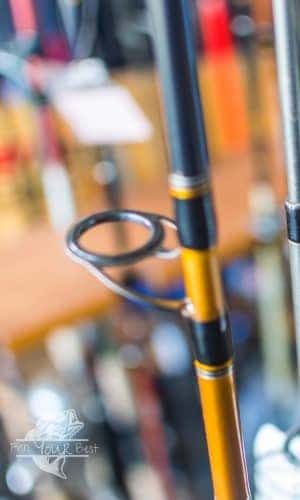 A close-up image of a fishing rod, focusing on one of its line guides with a black frame and ring. The rod has a glossy finish with gold trim accents, giving it a sleek appearance. The blurred background suggests a cluttered space, possibly a storage area or shop with various fishing gear. This detail shot highlights the important features of the rod's construction.