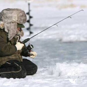 An individual bundled in heavy winter clothing, including a fur-lined hat and gloves, is seated on a frozen lake for ice fishing. They are holding a short ice fishing rod, attentively aiming it into a fishing hole cut into the ice. In the background, an ice auger stands upright, indicating the preparation work for the fishing spot. The environment appears cold and snowy, with a clear sky.