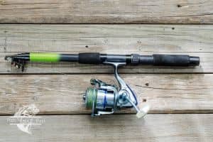 A telescopic fishing rod with a green tip and black foam handle lies on a wooden dock. A silver spinning reel is attached. The wooden planks provide a rustic background that contrasts with the modern fishing equipment, indicating the rod is set for use or has just been used.