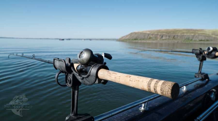 A close-up view of a trolling fishing rod mounted on the side of a boat. The rod is positioned horizontally, with its handle featuring a cork grip and the reel perched securely in a rod holder. The backdrop showcases a calm, expansive body of water with gentle ripples and a distant shore with low hills under a clear blue sky, indicating a serene day for fishing on the water.