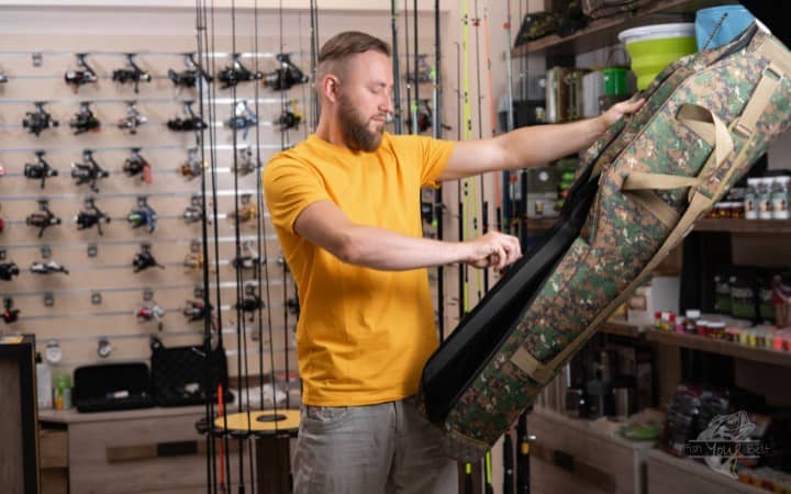 A man in a yellow t-shirt is inspecting a camouflage-patterned fishing rod carrying case in a well-organized fishing store. Behind him, an array of fishing rods with reels attached are displayed vertically on the wall, and various fishing gear products can be seen in the background. The man appears to be evaluating the case, possibly considering a purchase.