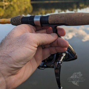 A close-up view of a person's hand gripping a fishing rod with a cork handle and a black spinning reel attached to it via a plastic reel seat. In the background, there is a blurred view of a calm lake during what appears to be the early evening, suggesting a peaceful fishing setting.