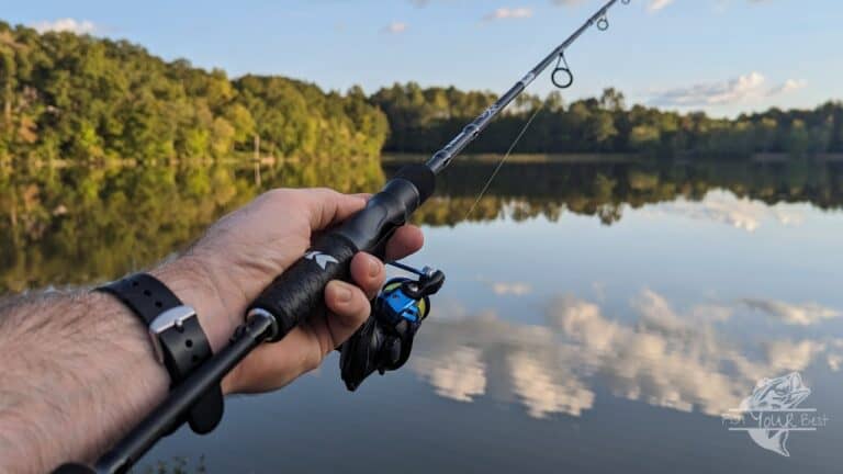 A close-up view of a person's hand holding a fishing rod, poised for casting, with a focus on the reel which is blue and black in color. The background features a tranquil lake with a forested shoreline, reflecting the trees and clouds in the calm water, suggesting an ideal fishing spot during a serene evening.
