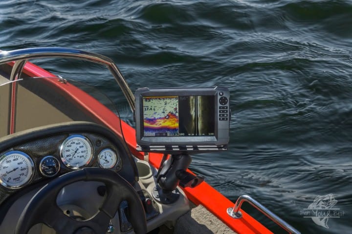 The image captures the dashboard of a fishing boat, equipped with a modern sonar fish finder displaying vivid colors that indicate underwater structures and fish. The screen shows a split view, with one side depicting the underwater topography and the other providing a detailed look that suggests the presence of fish, possibly crappie. In front of the sonar, the steering wheel of the boat is visible, alongside various gauges that monitor the boat's performance. The boat's vivid red edge stands out against the rippling dark blue waters of the lake, suggesting a clear day ideal for fishing.