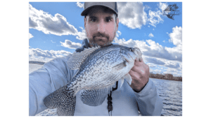 The image features a man engaged in winter crappie fishing on a lake. He is holding a large crappie close to the camera, showcasing its size and the successful catch. The man is dressed in a light-colored jacket suitable for cooler weather and wears a baseball cap. Behind him, the vast expanse of the main lake is visible with ripples of water reflecting the sky. The backdrop is a picturesque scene with blue skies, fluffy clouds, and a distant tree line, conveying a serene day of fishing in a winter setting.