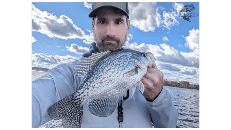 The image features a man engaged in winter crappie fishing on a lake. He is holding a large crappie close to the camera, showcasing its size and the successful catch. The man is dressed in a light-colored jacket suitable for cooler weather and wears a baseball cap. Behind him, the vast expanse of the main lake is visible with ripples of water reflecting the sky. The backdrop is a picturesque scene with blue skies, fluffy clouds, and a distant tree line, conveying a serene day of fishing in a winter setting.