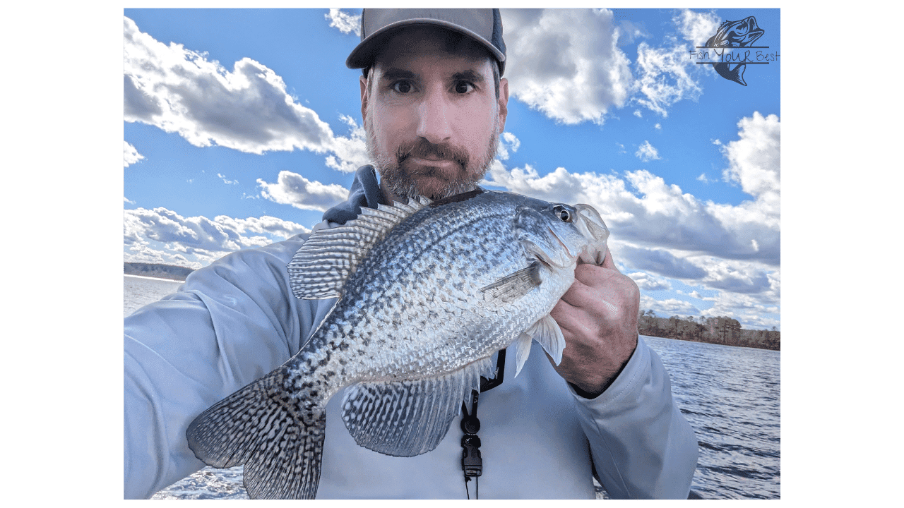 The image features a man engaged in winter crappie fishing on a lake. He is holding a large crappie close to the camera, showcasing its size and the successful catch. The man is dressed in a light-colored jacket suitable for cooler weather and wears a baseball cap. Behind him, the vast expanse of the main lake is visible with ripples of water reflecting the sky. The backdrop is a picturesque scene with blue skies, fluffy clouds, and a distant tree line, conveying a serene day of fishing in a winter setting.